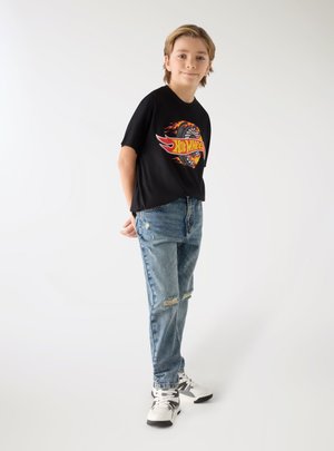 Młody chłopiec stoi z rękami za plecami, ubrany w czarną koszulkę Hot Wheels, podarte niebieskie jeansy i białe trampki na białym tle.