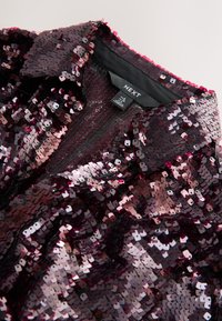 Burgundia värvi litritega top, millel on kõrge kaelus ja lühikesed varrukad. Iseloomulik tekstureeritud pind, kus on tihedalt paigutatud peegeldavad litrid. Brändi silt on nähtav.