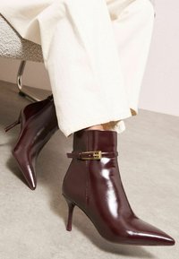 Burgundijas krāsas patentēta ādas potīšu zābaki ar punktētu purngalu, plānu stiletto papēdi un dekoratīvu sprādzi potītes rajonā.