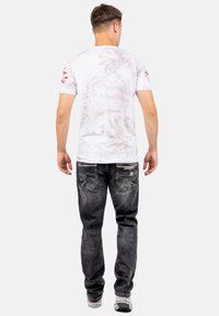 T-shirt bianca con texture marmorizzata, maniche corte e dettagli rossi sulle spalle; abbinata a jeans neri con tasche ricamate.
