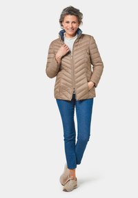 Giacca trapuntata beige con motivo a zigzag, colletto alto, due tasche laterali e chiusura con zip, abbinata a jeans blu e sneakers beige.