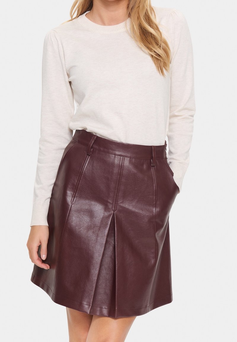 Femme portant un pull crème à manches longues rentré dans une jupe en cuir marron foncé taille haute avec un pli à l'avant et des poches.