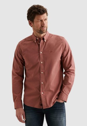 Mann mit braunen Haaren, der ein rostfarbenes Hemd mit Knöpfen und dunkle Jeans trägt, steht mit einer Hand in der Tasche und schaut seitlich.