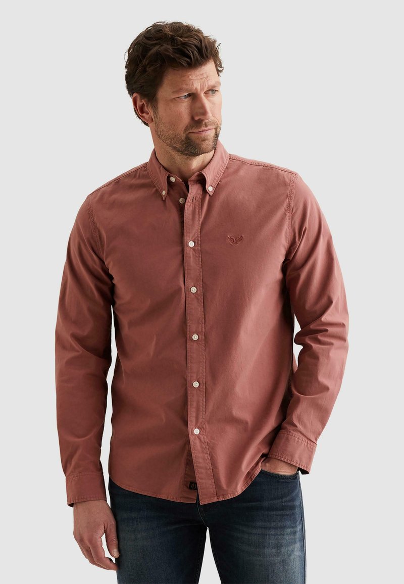 Muž s hnedými vlasmi v hnedej košeli na gombíky a tmavých džínsach, stojaci s jednou rukou v vrecku, pozerajúci sa do boku.