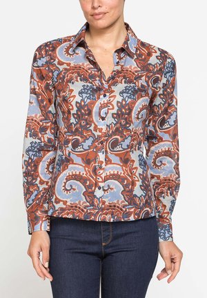 Donna che indossa una camicia a maniche lunghe abbottonata con un motivo paisley intricato nei colori ruggine, blu e bianco, abbinata a jeans blu scuro.