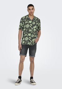 Kurzarm, dunkelgrünes Blusenshirt mit hellgrünen Mustern, kombiniert mit dunklen Jeansshorts und schwarzen Sneakern mit weißen Akzenten.
