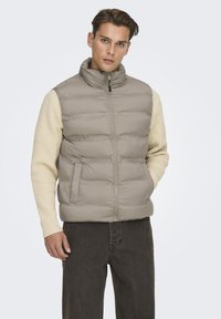 Gilet imbottito grigio chiaro con colletto alto, zip frontale e tasche laterali, indossato sopra un maglione a coste crema, abbinato a jeans scuri.