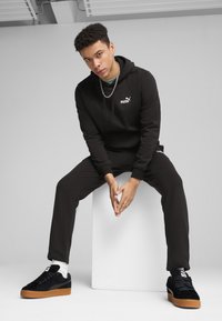 Hoodie noir avec logo, joggers assortis, et baskets en suède noir avec semelles en gomme. Les accessoires comprennent une chaîne en collier et des chaussettes blanches.