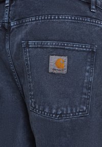 Farkut tummalla pesulla, joissa on takatasku ja neliönmuotoinen harmaa logo, jossa on oranssi tyylitelty "C".