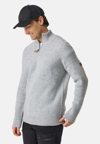 Man wearing a light gray ribbed sweater with a half-zip collar and black cap, smiling and looking down, against a plain background.