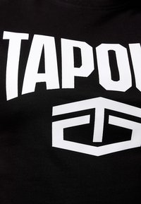 Melna kokvilnas t-kreklā ar drosmīgiem baltiem burtu blokiem, kas uzrakstīti "TAPOUT", un ģeometrisku logo, ar gludu tekstūru un noapaļotu apkakli.
