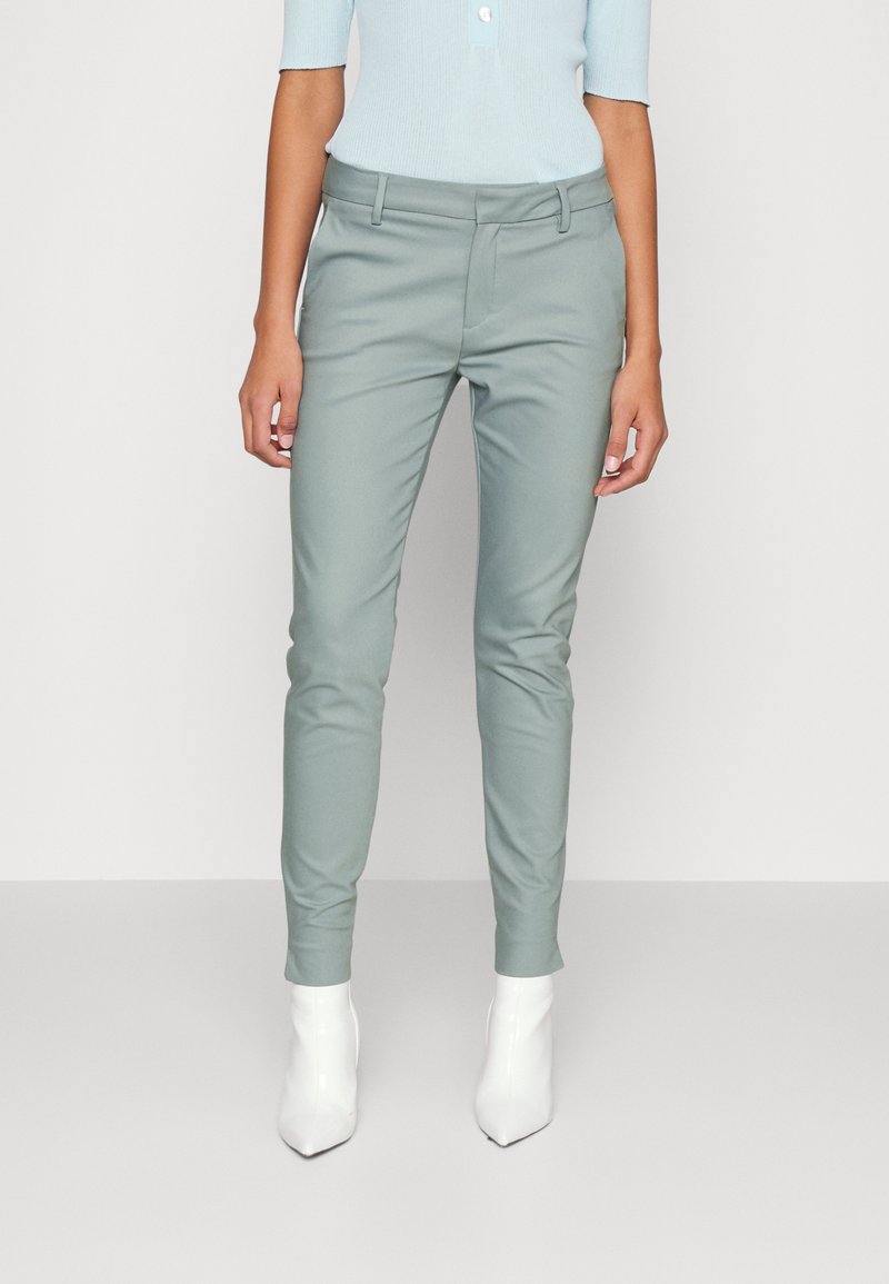 Femme portant un pantalon slim vert clair et des bottines blanches, debout devant un fond clair uni.