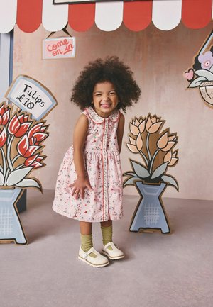 Mazā meitene smaidot, tērpusies rozā kleitiņā un zaļās zeķēs, stāv starp karikatūru stilā zīmētiem ziedu podiem zem sarkanbalta markīzes.