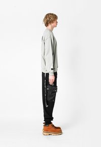 Sudadera gris claro, pantalones negros con bolsillo cargo y botas color beige. Tejido texturizado, corte relajado y acentos de logotipos visibles en las prendas.