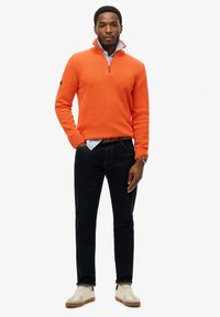 Oranger gestrickter Pullover mit einem Reißverschlusskragen, kombiniert mit dunklen Jeans und cremefarbenen Sneakers. Hinweis: lässiges Ensemble mit sichtbaren Texturen.
