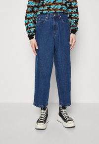 Calças largas de ganga azul com cintura alta, apresentando o clássico estilo de cinco bolsos e costuras visíveis. Combinadas com ténis pretos de cano alto.