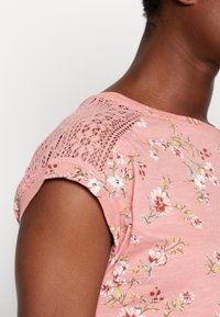 Lyserød blomstret t-shirt med korte ærmer, der har tekstureret blondeaccent på skulderen. Blødt, let stof. Blomsterdesign i hvid og blå.