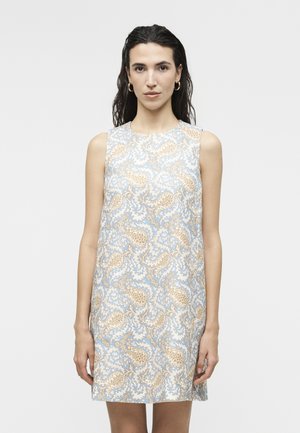 Γυναίκα που φοράει φόρεμα χωρίς μανίκια, μήκους μέχρι το γόνατο, με μοτίβο paisley σε μπλε και μπεζ αποχρώσεις, στέκεται μπροστά σε απλό ανοιχτό φόντο.