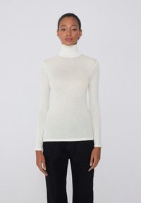Leichtes, enganliegendes Rollkragenshirt in Off-White, hergestellt aus einem weichen, dünnen Material mit langenÄrmeln und einer glatten Textur. Kombiniert mit schwarzen Hosen.
