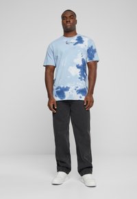 Světle modré tričko s tye-dye vzorem, tmavě modré vzory, bavlněný materiál, krátké rukávy a vyšívané logo nad levou hrudí.