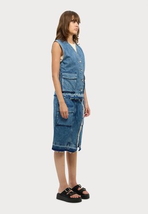 Gilet in denim con scollo a V, chiusura a bottoni e due tasche anteriori, abbinato a una gonna in denim lunga fino al ginocchio con orli sfrangiati. Sandali neri con zeppa.