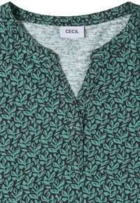 Haut en tissu vert foncé avec un motif de feuilles vert clair, encolure ronde fendue, et étiquette de marque CECIL visible à l'intérieur du col.