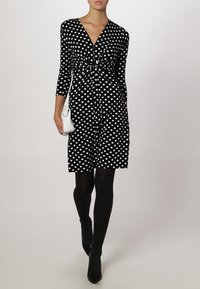 Svart klänning med vita prickar, v-ringning, trekvartslånga ärmar och figursydd midja. Bärs med svarta strumpbyxor och svarta klackar. Liten vit handväska.