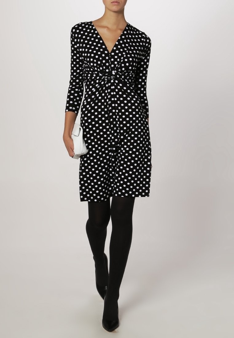 Svart klänning med vita prickar, v-ringning, trekvartslånga ärmar och figursydd midja. Bärs med svarta strumpbyxor och svarta klackar. Liten vit handväska.