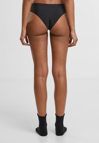 Zwarte hoge taille bikini onderzijde met een gladde textuur, minimale bedekking en een hoog uitgesneden beenontwerp, gecombineerd met zwarte geribbelde enkelkousen.
