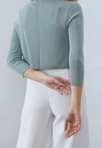 Mintgrön stickad topp med trekvartslånga ärmar, med en texturerad yta och en sömdetalj på baksidan, kombinerad med vita byxor.