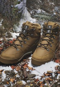 Brown hiking boots with black laces placed on a snowy forest floor with leaves, twigs, and tree bark in the background.