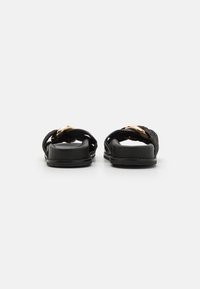 Black sandals with braided detailing, featuring a prominent gold-tone accent. The design is flat with a textured surface.