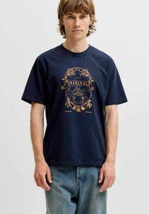 Młody mężczyzna o średniej długości brązowych włosach nosi granatowy T-shirt z pomarańczowym herbem w stylu vintage i niebieskie dżinsy, stojąc na jasnym tle.
