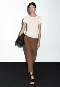 Vrouw die tegen een witte achtergrond loopt, gekleed in een wit T-shirt, een bruine rok, zwarte sandalen, en een grote zwarte gevlochten clutch bag vasthoudt.