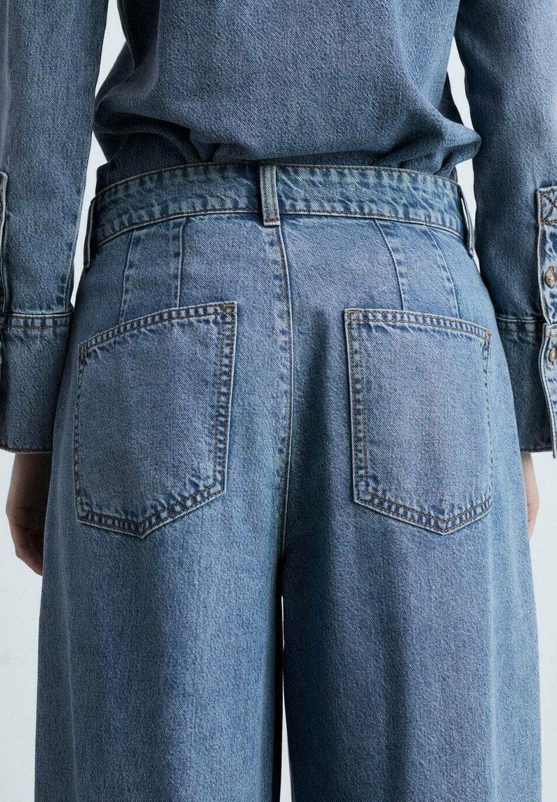 Vaaleansiniset farkut, joissa on löysä malli, kahdella takataskulla, näkyvillä ompelusaumojen yksityiskohdilla ja korkealla vyötärömallilla.