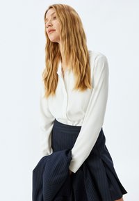 Blusa blanca con botones, de mangas largas; combinada con una falda de rayas en azul oscuro. Textura de tela suave, diseño minimalista, silueta elegante.