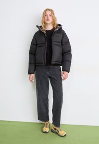 Svart boblejakke med hette, med glidelås foran og sømdetaljer. Kombinert med mørke jeans og gule joggesko med svarte detaljer.
