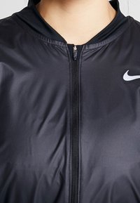 Veste noire zippée fabriquée en tissu lisse et léger. Dotée d'un col haut et d'un logo Nike blanc sur le côté gauche.