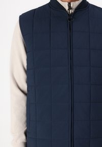 Marineblaues gesteppte Weste mit Reißverschluss und Stehkragen, mit glatter Textur und rechteckigen Mustern, getragen über einem hellen Pullover.