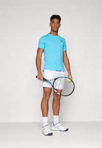 Hellblaues, kurzärmliges Sportshirt mit vertikalen Streifen, kombiniert mit weißen Shorts und weißen Turnschuhen. Hält einen Tennisschläger mit einem Ball.