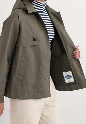 Osoba ubrana w oliwkowo-zieloną zapinaną na guziki kurtkę nałożoną na granatowo-biały pasiasty golf, trzymająca kurtkę otwartą, aby pokazać wewnętrzną metkę.