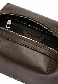 Sac en cuir marron à finition texturée, doté d'une fermeture éclair argentée et d'une doublure intérieure noire. Détail du logo visible à l'extérieur.