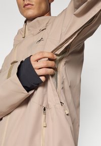 Beige dzseki magas gallérral és cipzáras nyitásokkal, hálós béléssel, állítható mandzsettákkal és ergonomikus varrásokkal a mozgás érdekében.