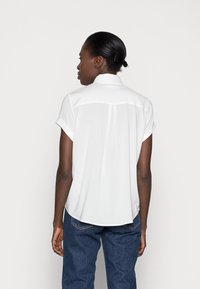 Blusa bianca a maniche corte con colletto, tessuto morbido, cucitura sul retro e orlo leggermente curva, abbinata a jeans in denim blu.