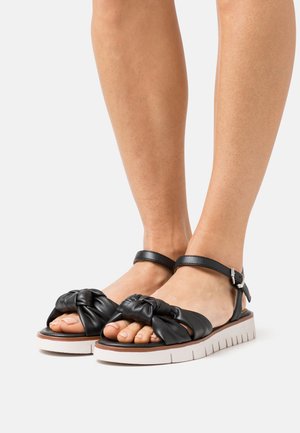 Sandalias de cuero negro con diseño anudado en las correas de los dedos, correa ajustable en el tobillo y suela blanca gruesa con textura.