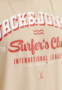 Béžové bavlněné tričko s odvážným červeným a bílým textem: "JACK&JONES Surfer's Club" s grafikou vln a kříženými čarami na spodní části.