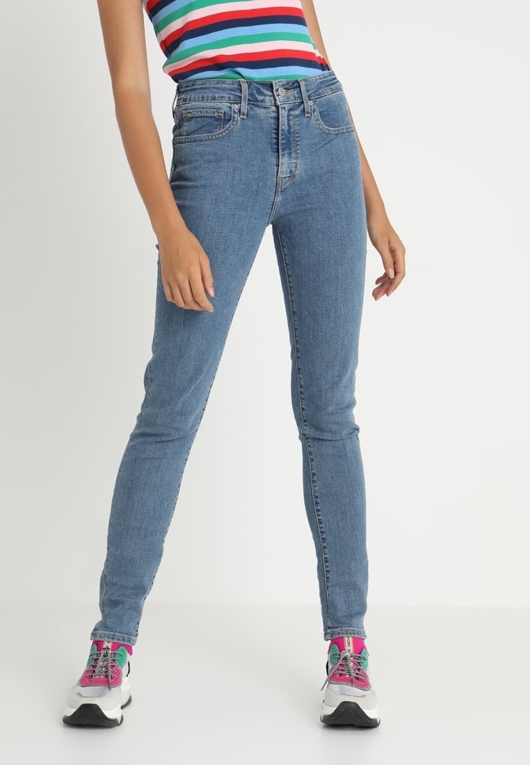Jeans ajustados azules de cintura alta con bolsillos frontales, combinados con una camiseta de tirantes a rayas multicolor y zapatillas deportivas coloridas.