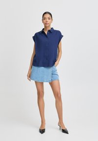 Camicia a maniche corte blu navy con bottoni abbinata a una gonna avvolgente in denim chiaro. Tacchi a punta neri completano l'outfit. Texture morbide e linee pulite.