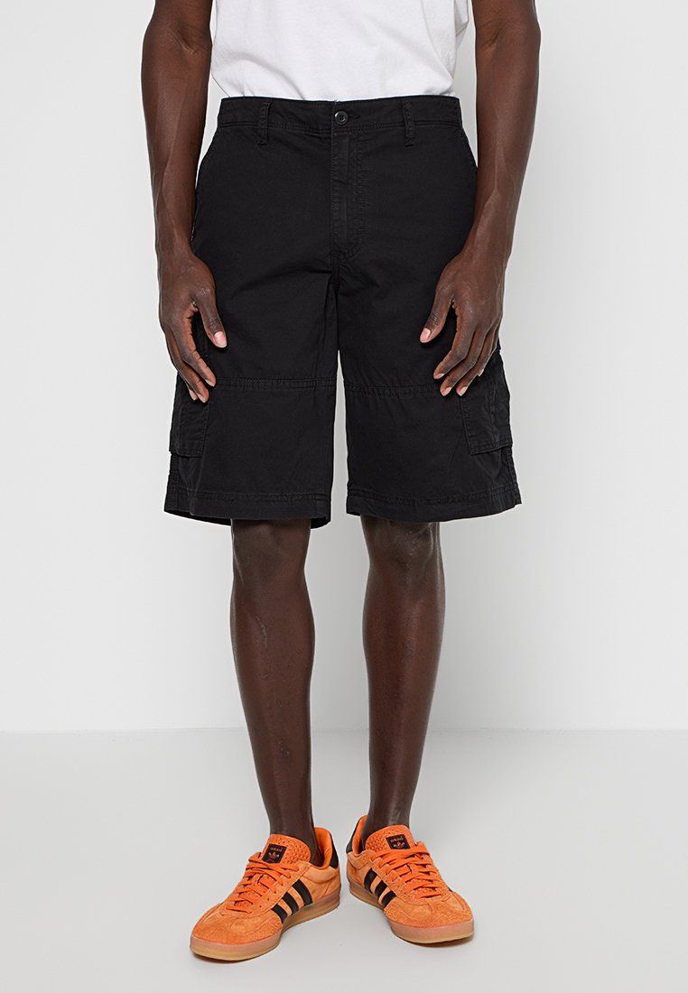 Schwarze Cargo-Shorts aus Baumwolle, mit Seitentaschen, geradem Schnitt und Knopfverschluss. Kombiniert mit orangefarbenen Turnschuhen.