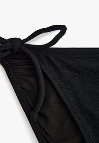 Czarny biustonosz bikini wykonany z fakturowanej tkaniny. Posiada szczegóły związane z wiązaniem ramiączka i gładkie krawędzie, podkreślające minimalistyczny styl.